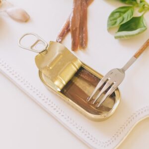Top view of an open can of anchovies with fork, basil, and garlic on white tray.