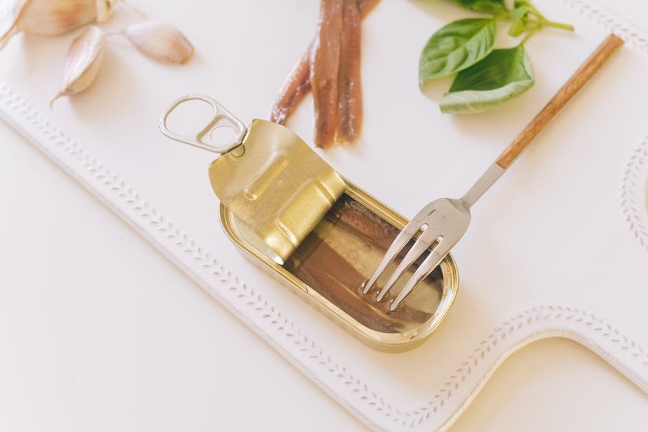 Top view of an open can of anchovies with fork, basil, and garlic on white tray.